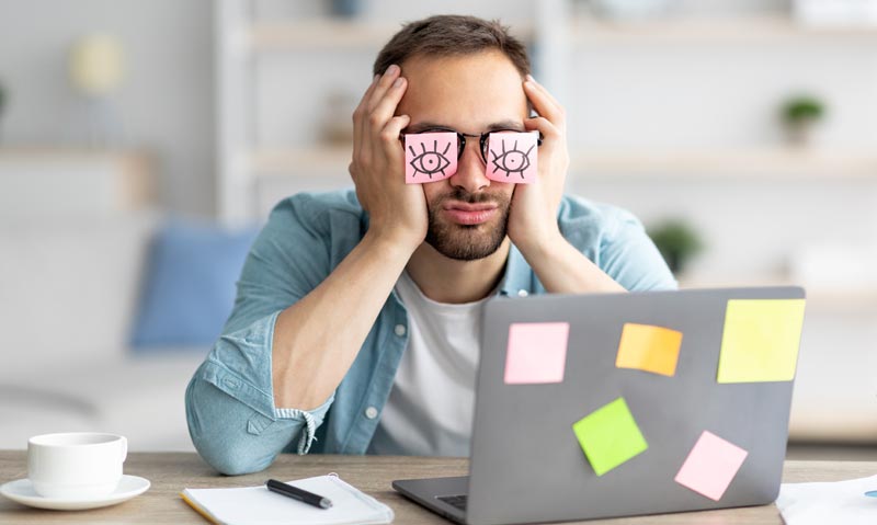 Übermüdeter Mann sitzt vor einem Laptop und hat Post-its mit aufgemalten Augen auf die Augen geklebt, Stichwort Chronische Insomnie.
(c) AdobeStock