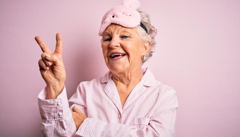 Fröhliche ältere Frau im Pyjama trägt eine Schlafmaske auf dem Kopf und zeigt ein Peace-Zeichen.
(c) AdobeStock
