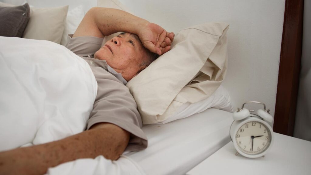 Ältere Person liegt nachts wach im Bett und blickt erschöpft zur Decke, neben ihr steht ein Wecker. (c) AdobeStock