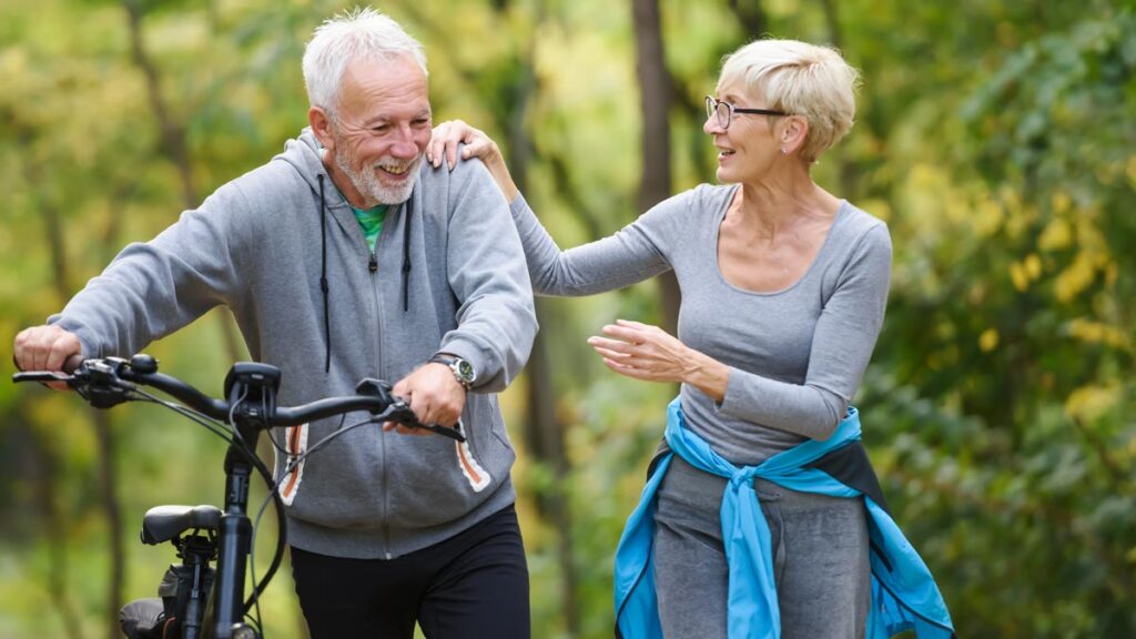 Ein lachendes Senioren-Paar schiebt ein Fahrrad durch einen frühlingshaften Wald und genießt das Frühlingserwachen. (c) AdobeStock