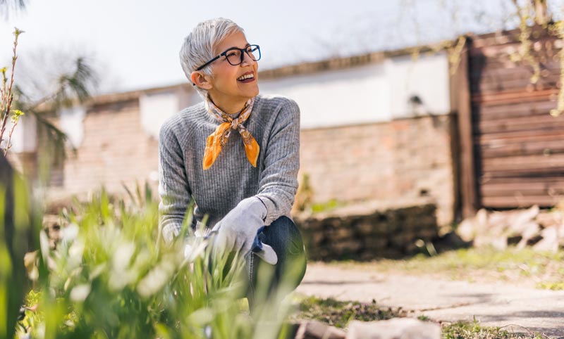 Eine lächelnde Frau mit kurzen grauen Haaren und gelbem Halstuch bei der Gartenarbeit im Frühling, Stichwort Frühlingserwachen.
(c) AdobeStock
