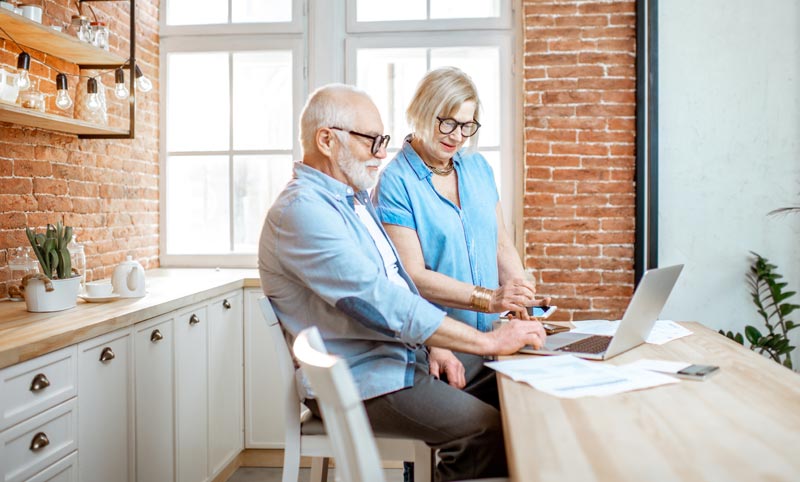 Älteres Paar sitzt in einer hellen Küche am Laptop und plant gemeinsam eine Reise am Computer.
(c) AdobeStock