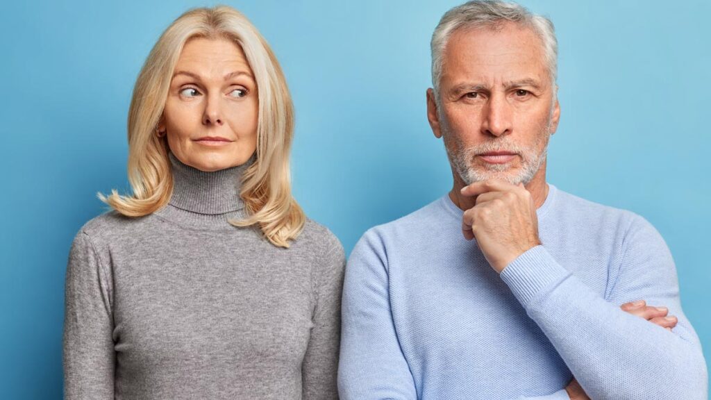Ein älteres Ehepaar steht vor einem blauen Hintergrund und grübelt über den Gender Life Gap nach. Die blonde Frau schaut nachdenklich zur Seite, während der graubärtige Mann sein Kinn hält. (c) AdobeStock