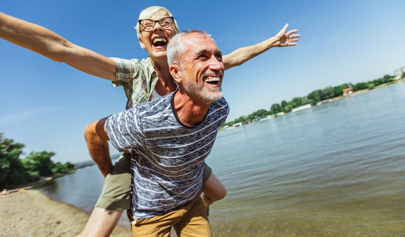 Ein glückliches älteres Paar genießt einen Sommertag am Wasser. Der Mann trägt die Frau im Huckepack, beide lachen ausgelassen und sie breitet die Arme aus. Die Sonne scheint, Stichwort Gender Life Gap.
(c) AdobeStock