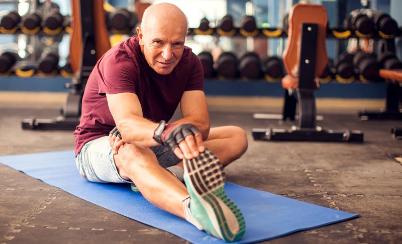 Ein fitter, kahlköpfiger älterer Mann im bordeauxroten T-Shirt sitzt auf einer blauen Matte im Fitnessstudio und dehnt sein Bein. Er schaut motiviert in die Kamera.
(c) AdobeStock