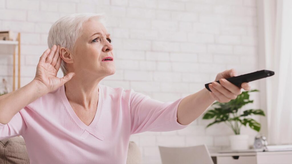 Eine ältere Frau sitzt auf einem Sofa, hält eine Fernbedienung und legt angestrengt die Hand ans Ohr, um besser zu hören. (c) AdobeStock