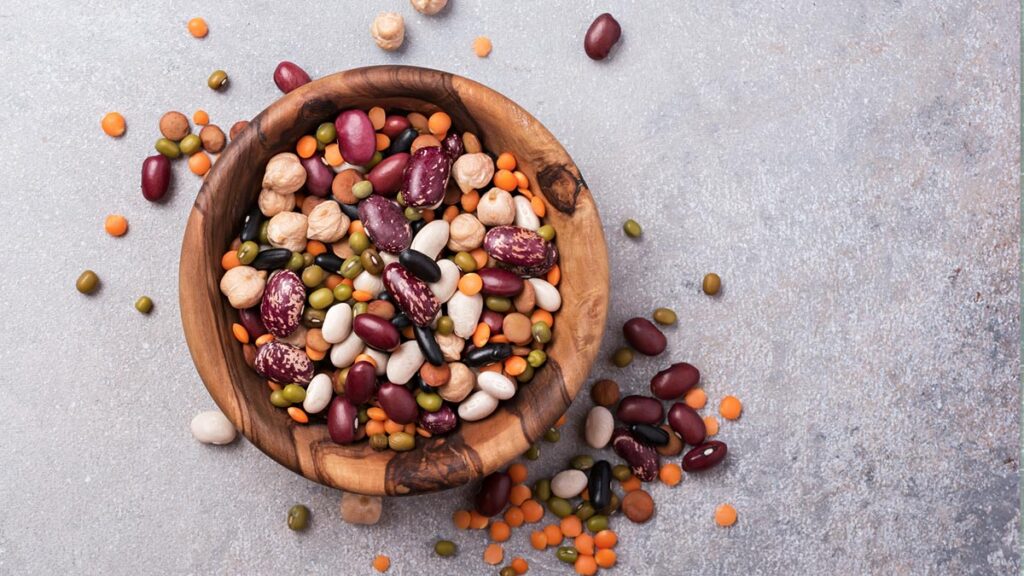 Eine Draufsicht auf eine rustikale Holzschale, gefüllt mit einer bunten Mischung aus verschiedenen getrockneten Hülsenfrüchten, darunter rote Kidneybohnen, weiße Bohnen, Kichererbsen, braune und rote Linsen sowie Mungobohnen. Die Schale steht auf einem grauen, strukturierten Steinhintergrund, mit einigen verstreuten Bohnen daneben. (c) AdobeStock