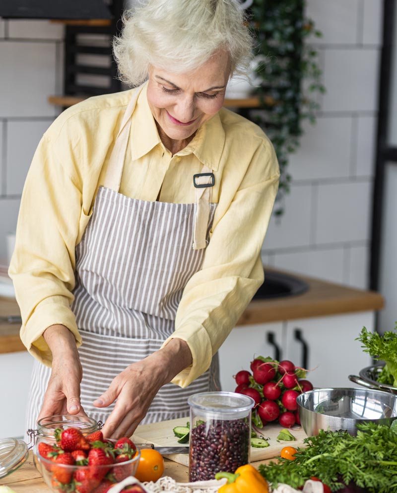 Eine lächelnde Seniorin mit grauen Haaren und einer gestreiften Schürze steht in einer hellen, modernen Küche und bereitet frisches Gemüse und Hülsenfrüchte zu. Auf der Arbeitsplatte liegen Erdbeeren, Paprika, Radieschen und ein Glas mit dunklen Bohnen, Stichwort Hülsenfrüchte.
(c) AdobeStock