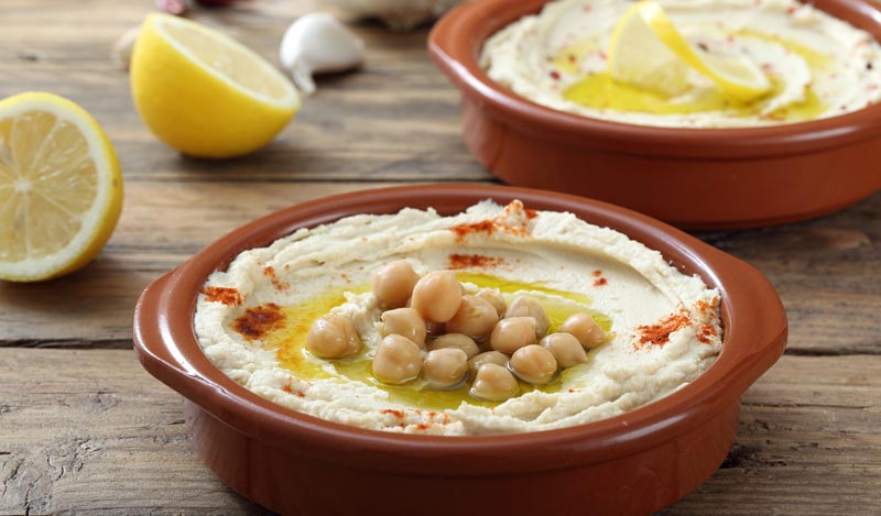 Zwei rustikale Terrakotta-Schalen mit cremigem Hummus stehen auf einem rustikalen Holztisch. Der vordere Hummus ist mit ganzen Kichererbsen, Olivenöl und rotem Paprikapulver garniert. Im Hintergrund sind frische Zitronenhälften und Knoblauchzehen zu sehen.
(c) AdobeStock