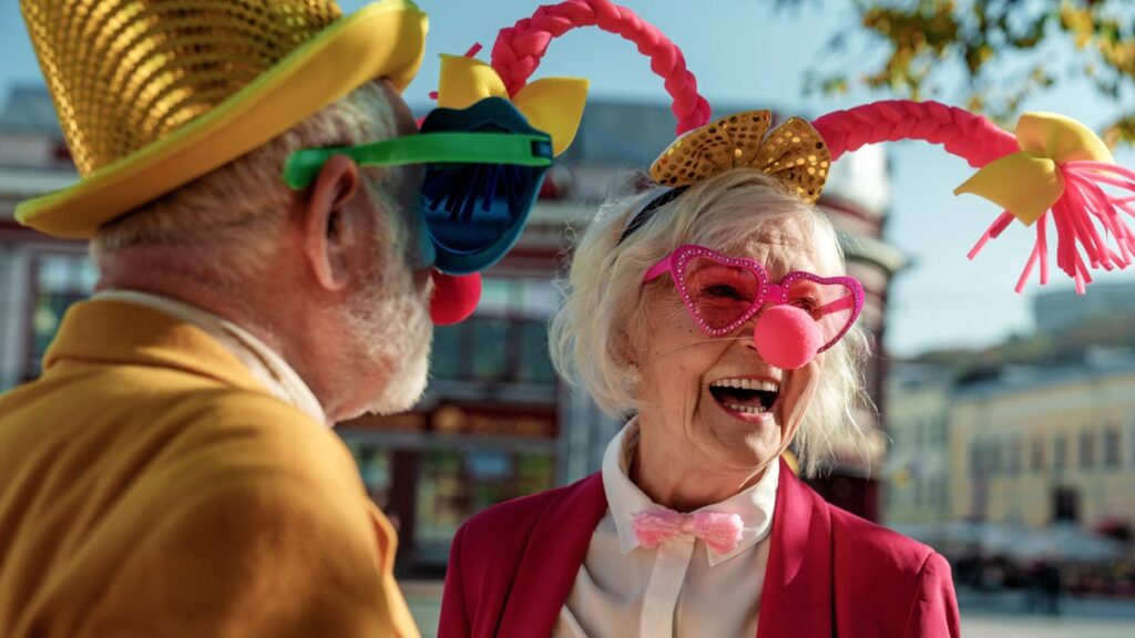Ein älteres Paar in farbenfroher Kleidung und albernen Kostüm-Accessoires lacht herzlich im Freien. Der Mann trägt einen goldenen Hut, die Frau eine pinke Perücke mit Antennen, eine Herz-Brille und eine Clownsnase. (c) AdobeStock
