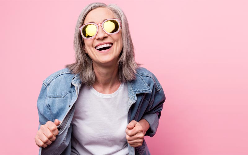 Eine Frau mit grauen Haaren, moderner Jeansjacke und gelb verspiegelter Sonnenbrille lacht ausgelassen vor einem pinken Hintergrund, Stichwort Humor und Gelassenheit.
(c) AdobeStock