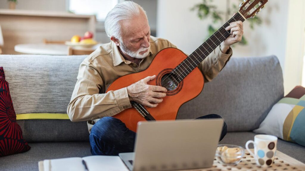 Ein Senior mit weißem Bart und Brille sitzt auf einem Sofa und lernt konzentriert, eine akustische Gitarre zu spielen. Vor ihm auf einem Couchtisch stehen ein offener Laptop, ein Notizbuch und ein Stift. (c) AdobeStock
