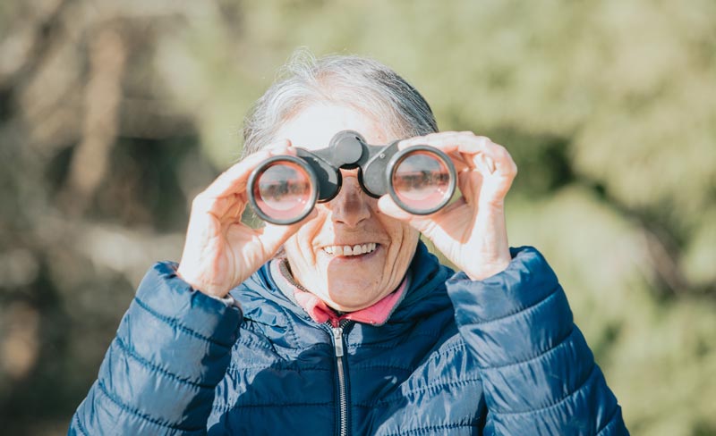Porträt einer lächelnden Seniorin mit grauen Haaren und blauer Daunenjacke, die im Freien ein schwarzes Fernglas an ihre Augen hält und hindurchschaut.
(c) AdobeStock
