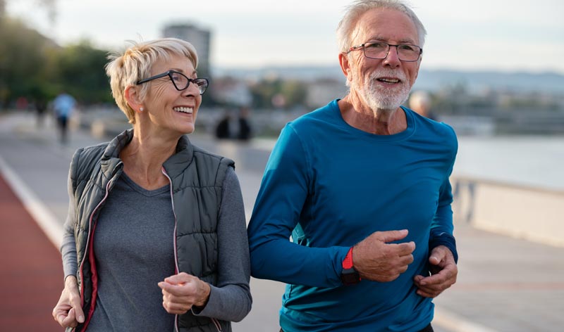 Ein sportliches älteres Paar joggt lächelnd im Freien. Der Mann trägt ein blaues Funktionsshirt, die Frau eine graue Weste über einem Longsleeve. Beide strahlen Vitalität und Freude an der Bewegung aus.
(c) AdobeStock