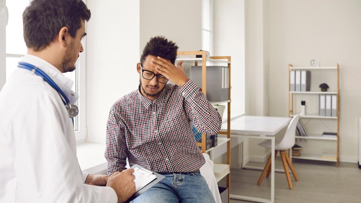 Die ultimative Migräne-Checkliste: Ihr Wegweiser aus dem Schmerzlabyrinth Ein Patient mit Kopfschmerzen im Gespräch mit einem Arzt im weißen Kittel in einer hellen Praxis. Der Arzt hält ein Klemmbrett. Fokus: Ärztliche Beratung und Migräne-Diagnose. (c) AdobeStock