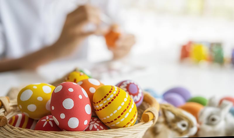 Ein Weidenkorb gefüllt mit bunt bemalten Ostereiern (Punkte, Streifen, Muster) steht im Vordergrund auf einem Tisch. Im unscharfen Hintergrund sind die Hände einer Person zu sehen, die gerade ein Ei bemalt, sowie ein Hase und weitere Eier, Stichwort Osterdeko selbst machen.
(c) AdobeStock