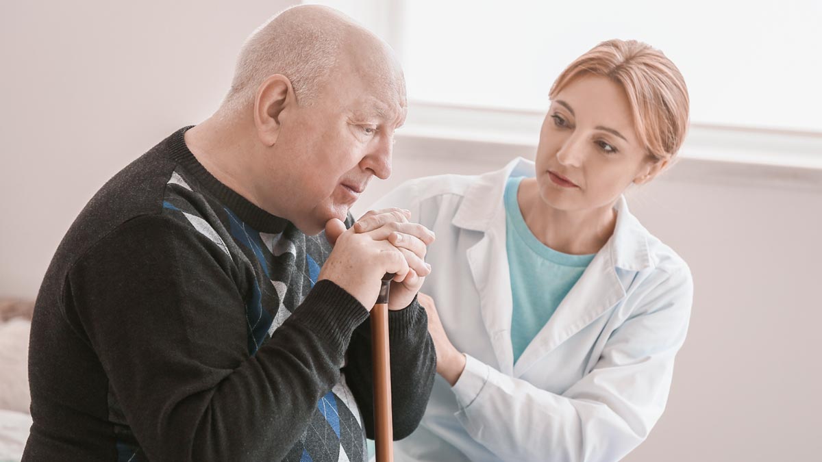 Parkinson Therapie in Österreich: Der stille Durchbruch gegen den Kontrollverlust Älterer Mann mit Parkinson sitzt nachdenklich mit Gehstock, eine Ärztin in weißem Mantel steht unterstützend hinter ihm in einem hellen Raum. (c) AdobeStock