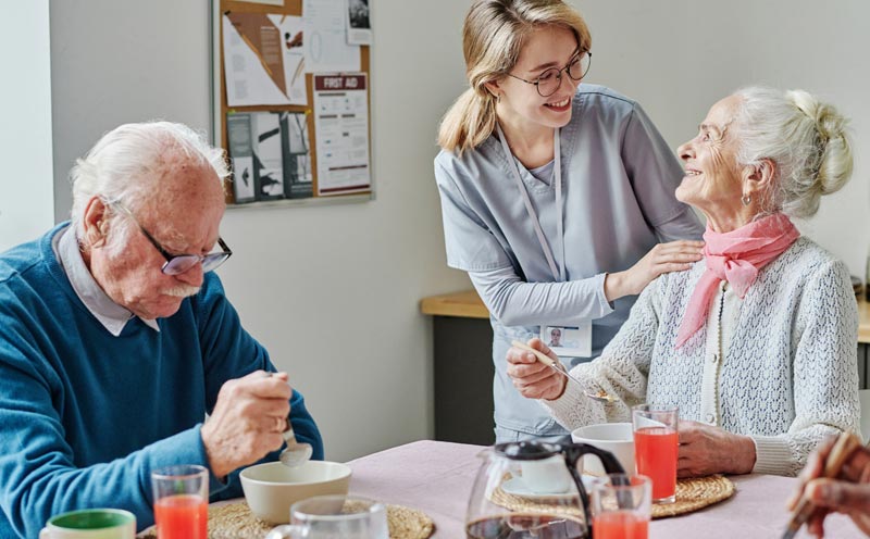 Ein Blick auf einen Esstisch in einem Gemeinschaftsraum. Mehrere ältere Menschen sitzen am Tisch und frühstücken. Eine junge Pflegerin steht unterstützend hinter einer älteren Frau und legt ihr die Hand auf die Schulter.
(c) AdobeStock