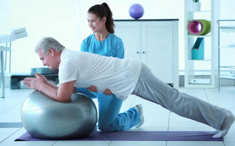 Eine junge Physiotherapeutin hilft einem älteren Mann, eine Plank-Übung auf einem Gymnastikball auszuführen, Stichwort Physikalische Medizin.
(c) AdobeStock