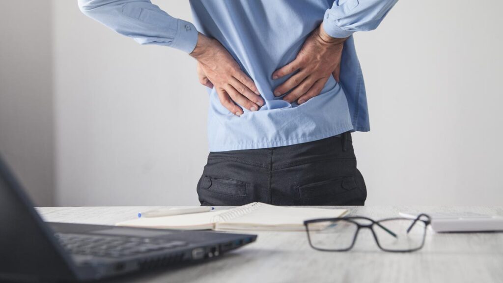 Ein Mann am Schreibtisch hält seine Hände an seinen unteren Rücken, um Schmerzen anzuzeigen. Im Vordergrund ein offener Laptop und eine Brille. (c) AdobeStock