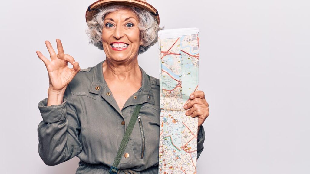 Porträt einer lachenden, älteren Frau mit lockigen grauen Haaren und Tropenhelm, die eine Karte hält und das OK-Zeichen macht. (c) AdobeStock