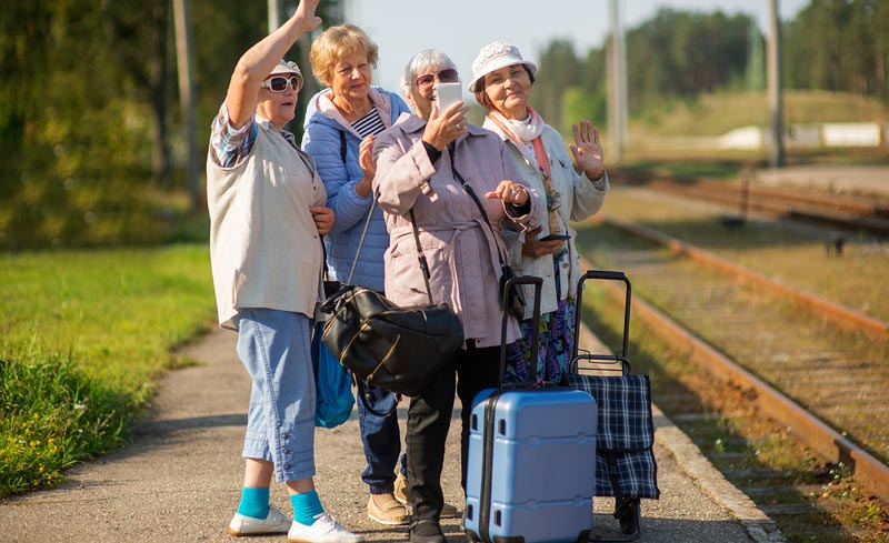 Vier ältere Frauen mit Reisekoffern und Handtaschen stehen auf einem ländlichen Bahnsteig, eine macht ein Selfie, eine andere winkt.
(c) AdobeStock