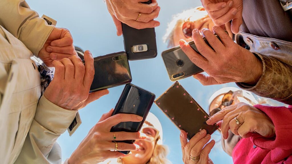 Mehrere Hände von Senioren halten ihre Smartphones in einem Kreis, fotografiert aus einer extremen Froschperspektive gegen einen blauen Himmel. (c) AdobeStock