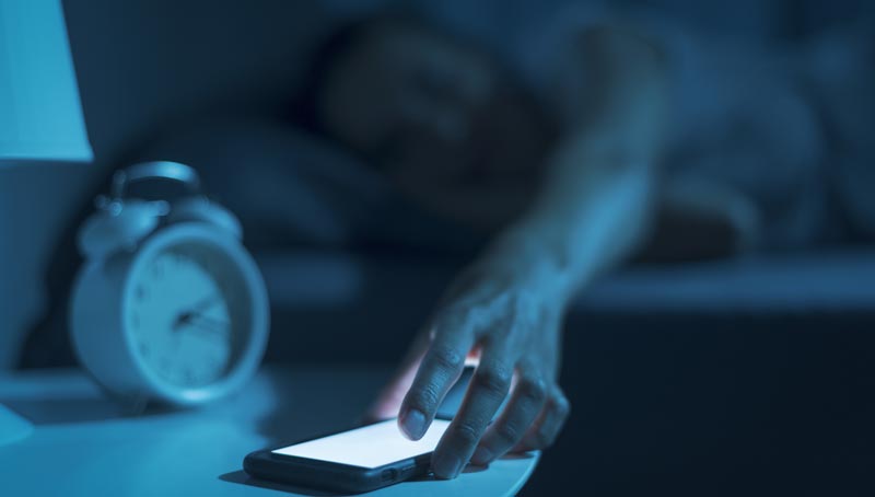in düsteres Schlafzimmer im blauen Licht eines Nachttisches und eines Weckers. Eine ausgestreckte Hand greift nach einem leuchtenden Smartphone auf dem Tisch, im Hintergrund liegt eine unscharfe Person im Bett, Stichwort Social Media Sucht im Alter.
(c) AdobeStock
