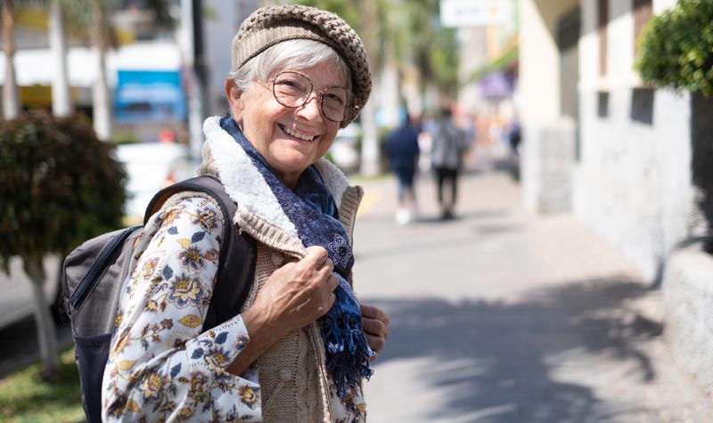 Eine lächelnde Seniorin mit grauen Haaren, Brille, Baskenmütze und einem Rucksack steht auf einer belebten Einkaufsstraße und blickt direkt in die Kamera.
(c) AdobeStock