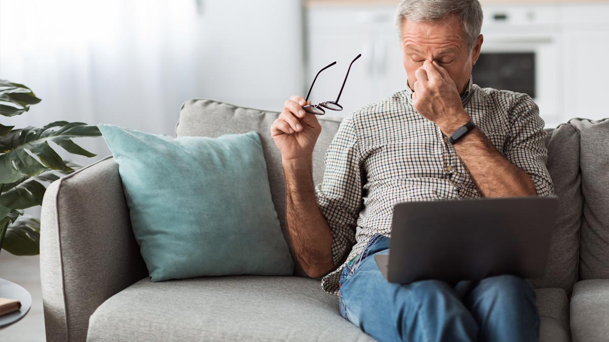 Vitaminmangel Symptome: Warum Sie trotz Schlaf ständig müde sind Älterer Mann mit Brille in der Hand sitzt erschöpft auf grauer Couch vor einem Laptop und reibt sich die Augenpartie. (c) AdobeStock