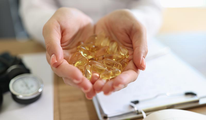 Vitaminmangel Symptome: Warum Sie trotz Schlaf ständig müde sind 2 Nahaufnahme von zwei Frauenhänden, die eine Handvoll goldgelbe, durchscheinende Fischöl-Kapseln (oder ähnliche Supplemente) präsentieren, im Hintergrund ein Schreibtisch mit medizinischen Unterlagen, Stichwort Vitaminmangel Symptome.
(c) AdobeStock