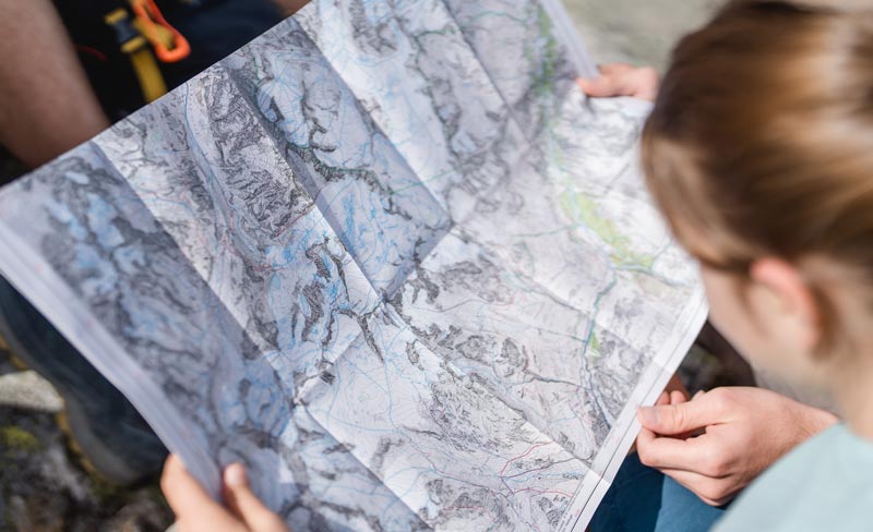 Die Draufsicht auf zwei Personen, die gemeinsam eine ausgebreitete topografische Wanderkarte aus Papier studieren. Die Hände halten die Karte fest, im Hintergrund ist vage alpine Ausrüstung und felsiger Untergrund zu erkennen.
(c) Alpennverein/ Sissi Richter