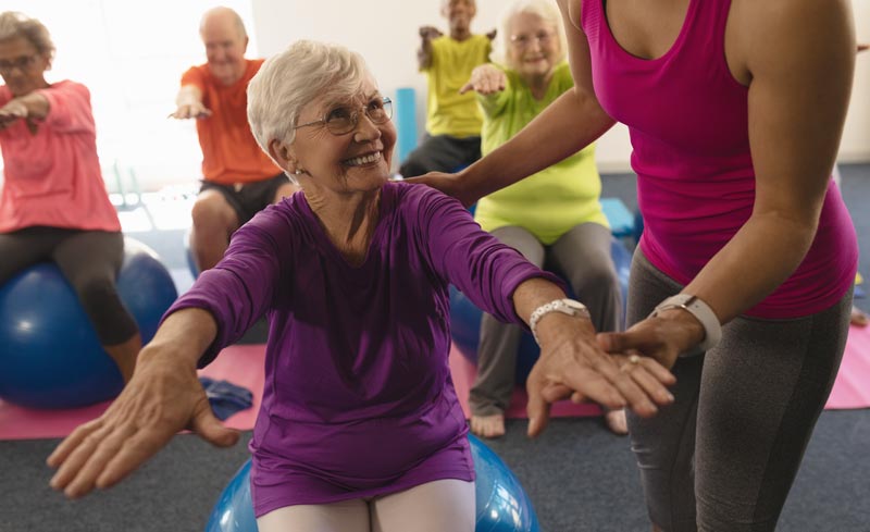 Eine ältere Frau mit Brille sitzt auf einem blauen Gymnastikball und wird von einer Trainerin in einem lila Tanktop angeleitet, während im Hintergrund eine Gruppe von Senioren trainiert.
(c) AdobeStock