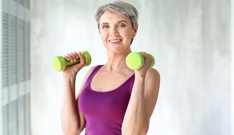 Eine ältere Frau mit kurzen grauen Haaren und einem lila Tanktop lächelt direkt in die Kamera, während sie zwei grüne Hanteln auf Schulterhöhe hält, Stichwort Bewegung im Alter.
(c) AdobeStock