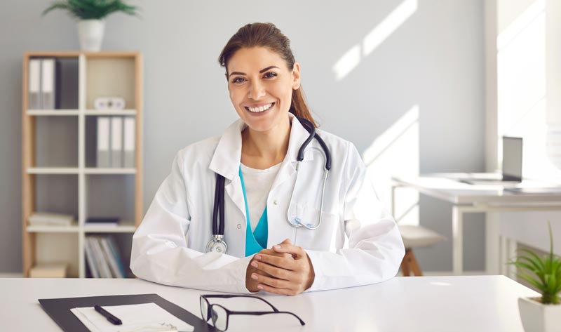 Eine freundlich lächelnde Ärztin mit braunem Haar und weißem Arztkittel sitzt an einem Schreibtisch und blickt direkt in die Kamera. Sie hat ihre Hände vor sich verschränkt. Ein Stethoskop hängt um ihren Hals. Im Hintergrund ist ein modernes, helles Büro mit einem Holzregal, Ordnern und einer Topfpflanze zu sehen. Sonnenlicht fällt durch ein Fenster, Stichwort Gesundheitswesen.
(c) AdobeStock