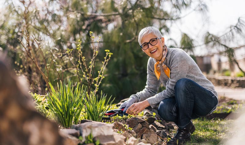 Seniorin kniet im Garten und jätet Unkraut. Sie trägt Handschuhe, eine Brille und ein Halstuch. Sie lächelt in die Kamera. Im Hintergrund sind blühende Pflanzen und ein Baum zu sehen.
(c) AdobeStock