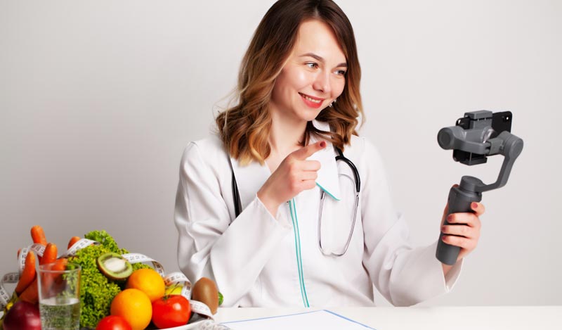 Eine lächelnde Ernährungsberaterin im weißen Arztkittel sitzt an einem Schreibtisch mit Obst, Gemüse und Vitamin-Ergänzungsmitteln und zeigt auf ein Smartphone, das auf einem Gimbal-Stativ montiert ist und live streamt, Stichwort Health Influencer.
(c) AdobeStock