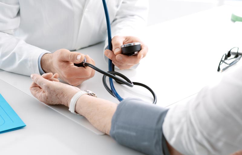 Ein Arzt im weißen Kittel misst den Blutdruck einer älteren Patientin am Handgelenk mit einer Blutdruckmanschette und einem Stethoskop. Ein Brillen-Etui und ein blauer Ordner liegen auf dem Tisch.
(c) AdobeStock