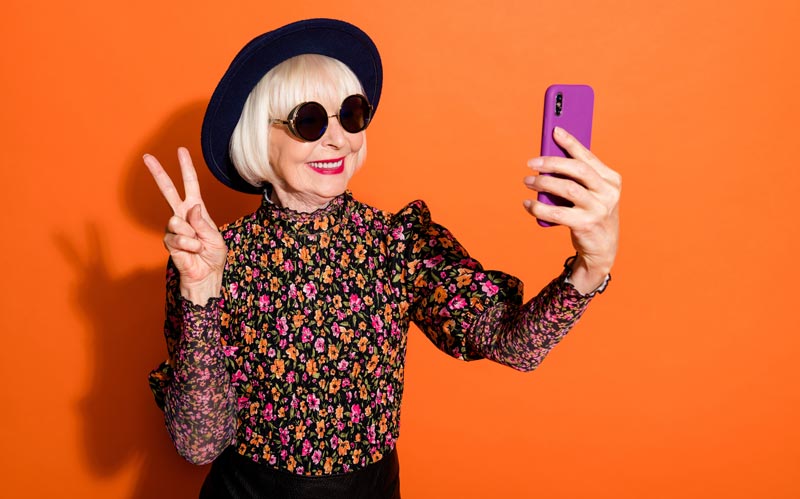 Stylische ältere Frau mit weißem Bob, Sonnenbrille und Hut macht ein Selfie mit einem lilafarbenen Smartphone und zeigt das Peace-Zeichen vor orangefarbenem Hintergrund, Stichwort Hochaltrigenstudie 2026.
(c) AdobeStock