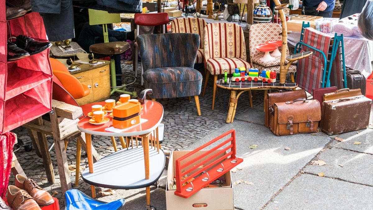 Keller entrümpeln: Der ultimative Guide, um 40 Jahre ‚Altlasten‘ diskret zu entsorgen Ein gut organisierter Flohmarktstand mit gut erhaltenen Mid-Century-Möbeln, darunter ein gemusterter Sessel, zwei gestreifte Stühle, ein rot-grauer Servierwagen und braune Lederkoffer, die ordentlich auf Steinplatten arrangiert sind. (c) AdobeStock