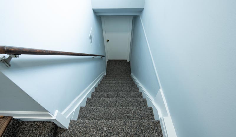 Keller entrümpeln: Der ultimative Guide, um 40 Jahre 'Altlasten' diskret zu entsorgen 1 Eine steile, mit dunklem Teppich ausgelegte Kellertreppe, aufgenommen aus der Vogelperspektive, die nach unten zu einer geschlossenen weißen Tür in einem hellblau gestrichenen Flur führt, Stichwort Keller entrümpeln.
(c) AdobeStock