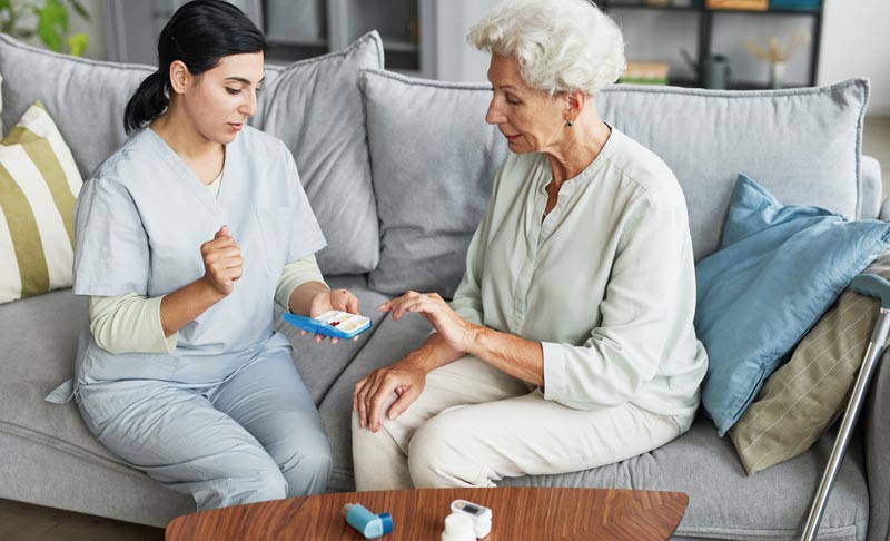 Eine junge Pflegerin in blauer Arbeitskleidung sitzt auf einem Sofa und zeigt einer älteren Frau mit weißen Haaren einen Medikamenten-Organizer. Auf dem Tisch davor liegen Inhalator und Tablettenflaschen.
(c) AdobeStock