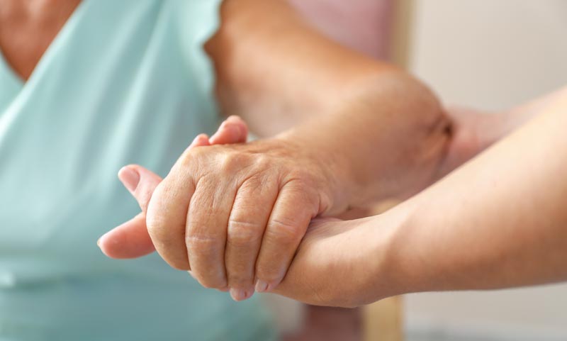 Eine Nahaufnahme, die zeigt, wie eine jüngere Hand die faltige Hand einer älteren Person stützend und tröstend hält. Im Hintergrund ist unscharf eine Person in hellgrüner Kleidung zu sehen, Stichwort pflegende Angehörige Hilfe.
(c) AdobeStock