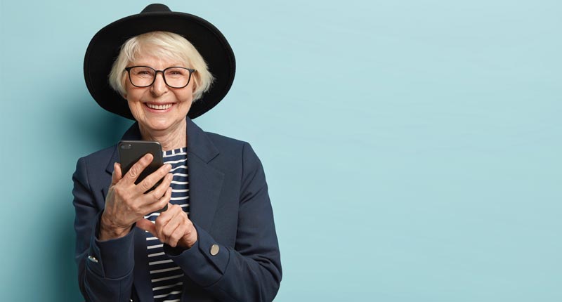 Eine stilvolle, ältere Frau mit weißem Kurzhaarschnitt, schwarzem Hut, Brille und einem dunkelblauen Blazer hält ein modernes Smartphone und lächelt wach in die Kamera. Der Hintergrund ist hellblau, Stichwort Seniorenhandy.
(c) AdobeStock