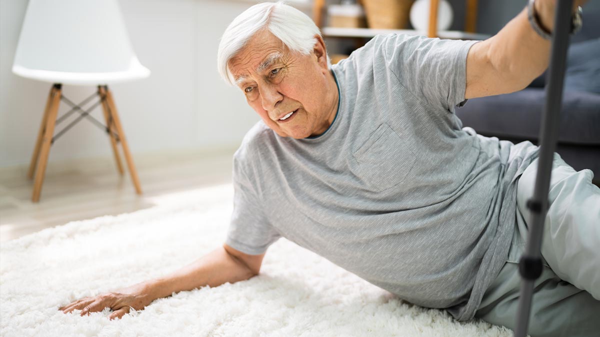 Sturzprävention im Alter: Warum ein sicherer Stand die beste Lebensversicherung ist Ein älterer Mann mit weißen Haaren stürzt in seinem Wohnzimmer auf einen weißen Teppich und versucht, sich an einer Haltestange oder Krücke festzuhalten. Er sieht verunsichert aus. (c) AdobeStock