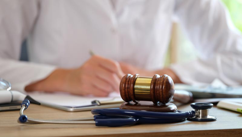 Ein hölzerner Richterhammer liegt neben einem Stethoskop auf einem Schreibtisch, im Hintergrund schreibt eine Person im weißen Arztkittel, Stichwort Wahlarzt.
(c) AdobeStock