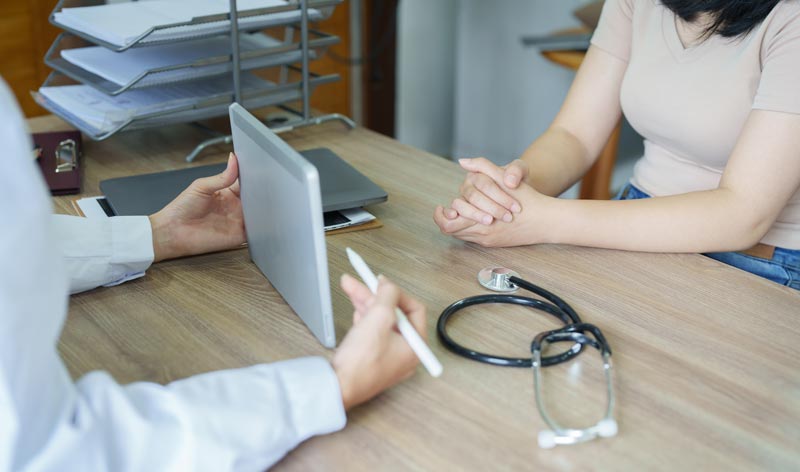 Eine Ärztin im weißen Kittel hält ein Tablet und erklärt einer Patientin, die ihre Hände gefaltet hat, medizinische Details an einem Holztisch.
(c) AdobeStock