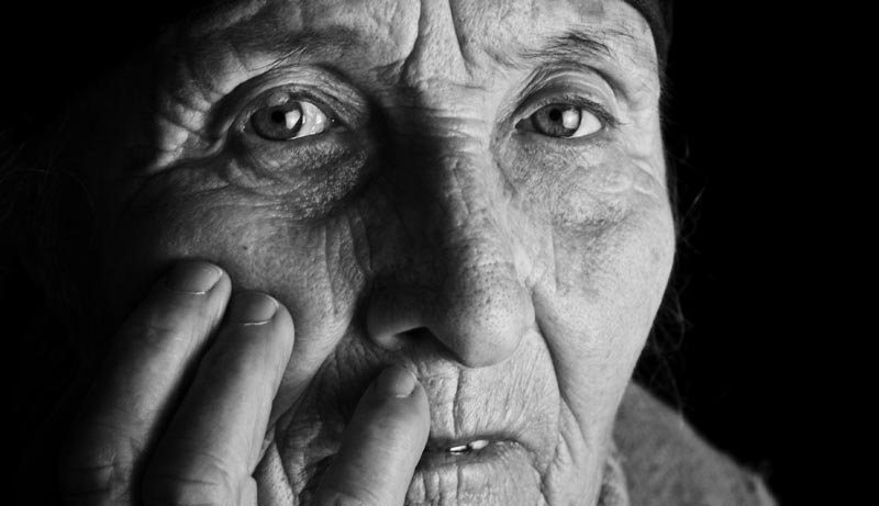 Extremes Schwarz-Weiß-Nahaufnahme eines älteren Gesichts. Jede Falte und Hautstruktur ist durch hartes Seitenlicht deutlich erkennbar. Der Blick ist direkt, wach und ernst. Die Hand berührt leicht die Wange, Stichwort Well-Aging.
(c) AdobeStock