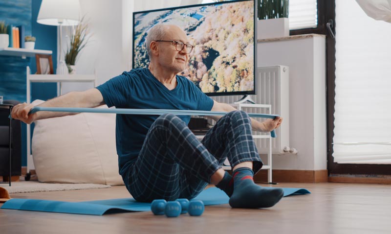 Ein älterer Mann mit Brille und Pyjamahose sitzt auf einer blauen Yogamatte im Wohnzimmer. Er dehnt ein blaues Widerstandsband mit beiden Händen vor seiner Brust. Im Vordergrund liegen kleine blaue Hanteln, im Hintergrund steht ein Fernseher.
(c) AdobeStock