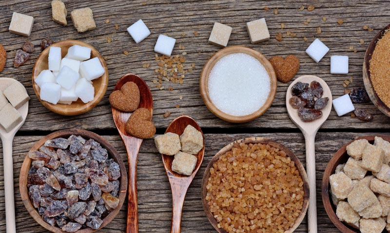 Draufsicht auf verschiedene Zuckersorten in Holzschalen und auf Löffeln auf rustikalem Holzuntergrund, darunter brauner Zucker, weißer Würfelzucker und Kandis.
(c) AdobeStock